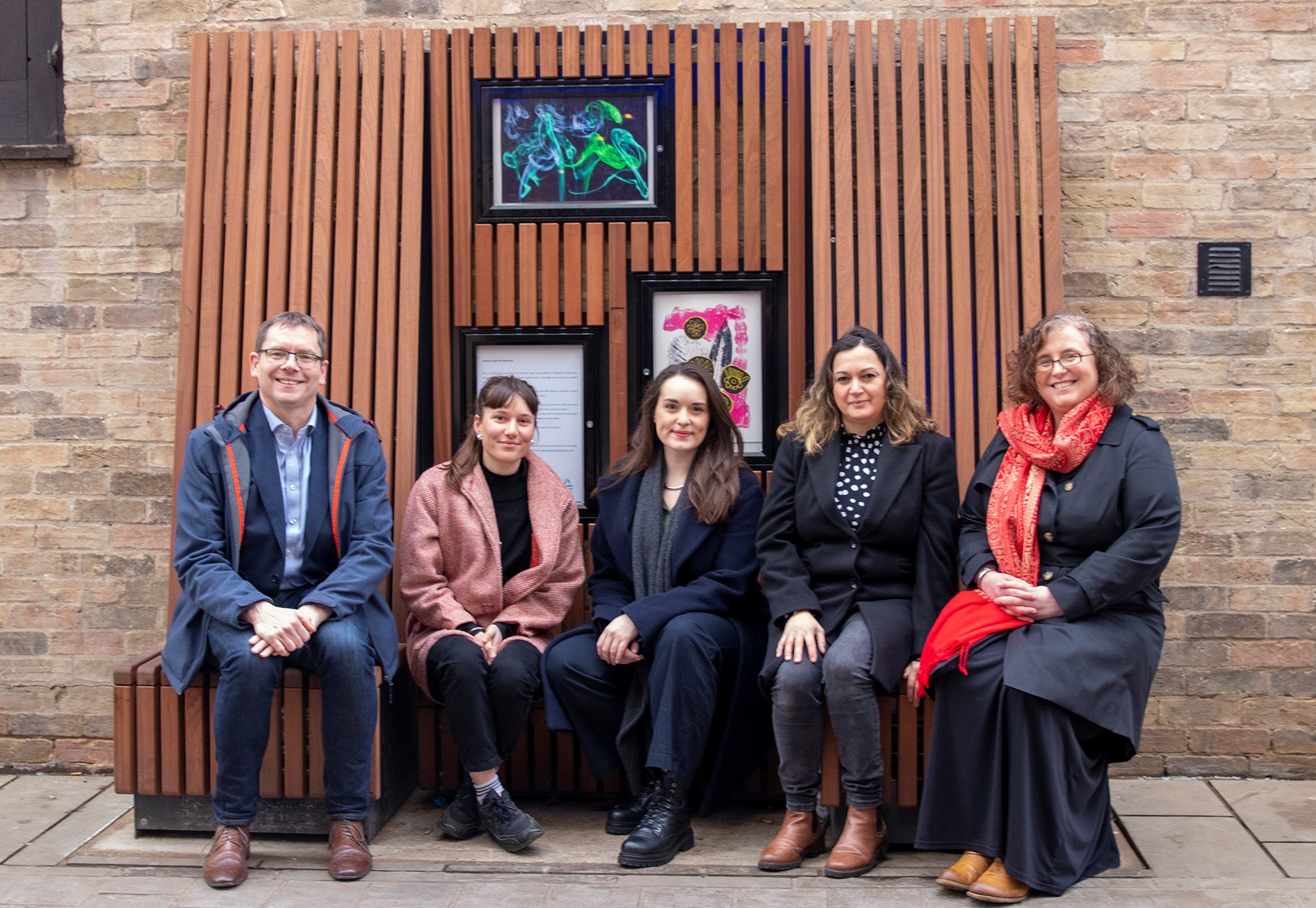 a man a four women sitting on a wooden bench outside