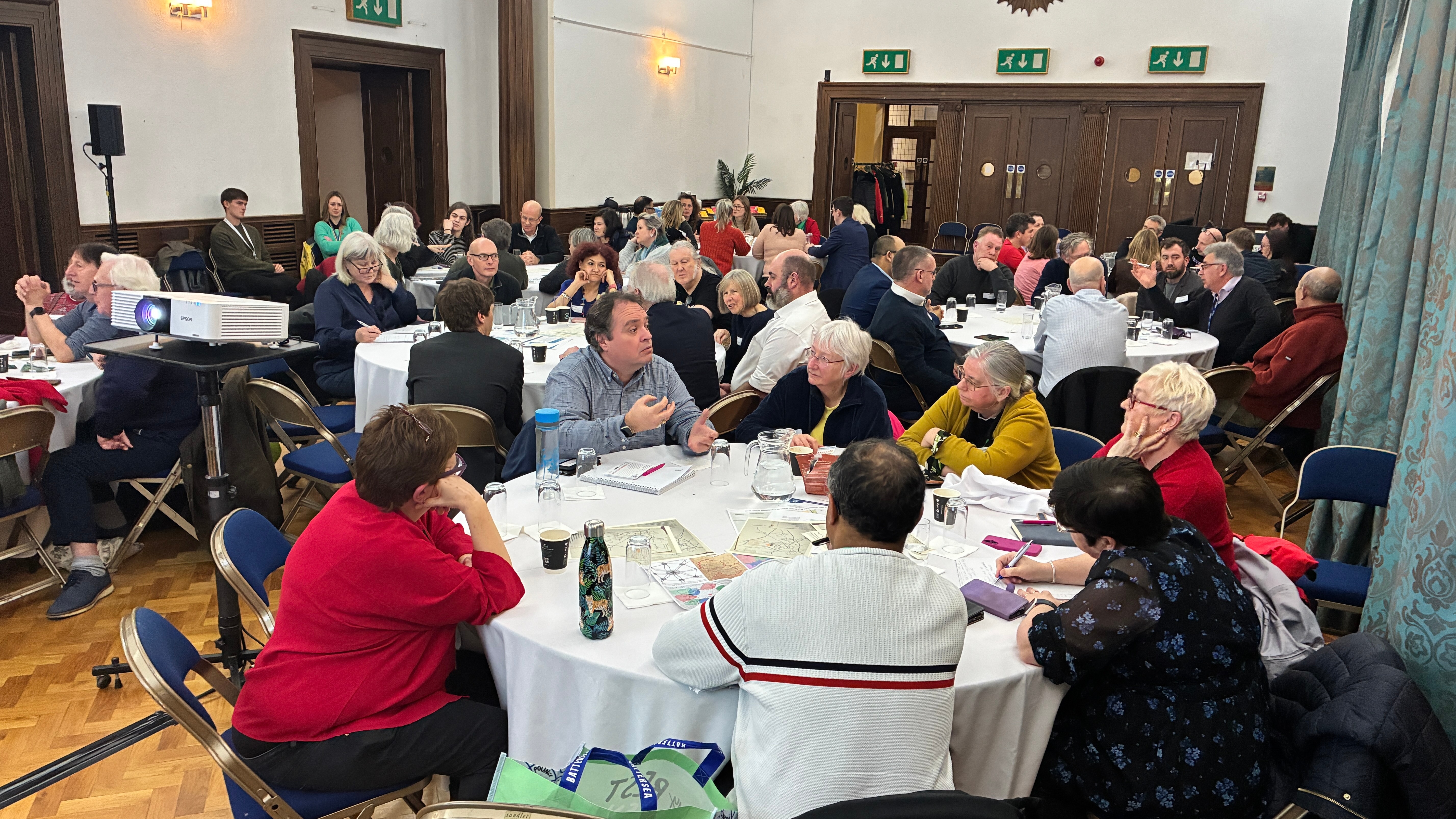 Large group of people seated around round tables in The Guildhall (Cambridge), engaged in discussion during a workshop-style event. Attendees are talking in small groups, with papers, maps and notebooks spread across the tables
