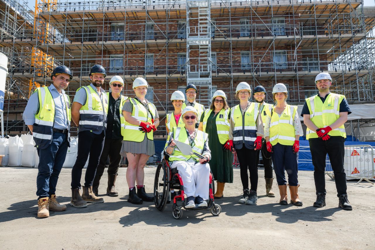 Lots of people standing in front of a building with scaffolding, with Cllr Gerri Bird in the centre holding a trowel